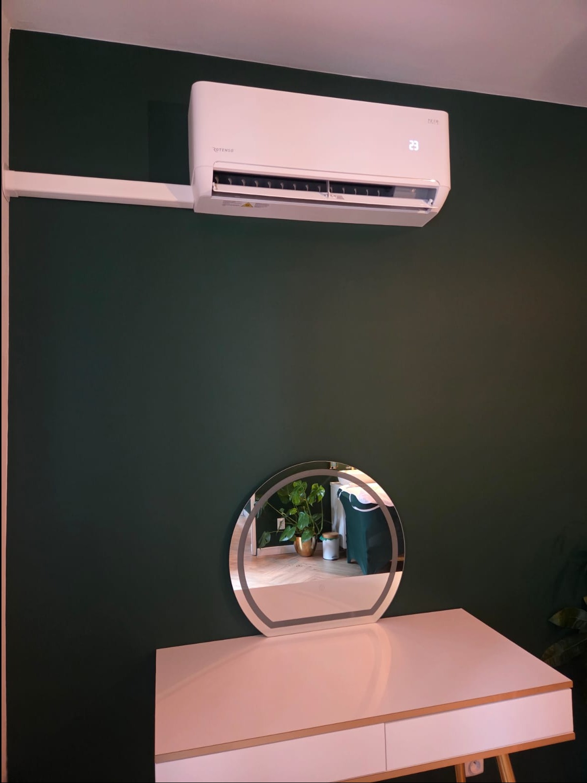 Biały klimatyzator Rotesno Teta 3.5 kW na zielonej ścianie nad toaletką z lustrem w salonie masażu. Widoczny wyświetlacz temperatury.