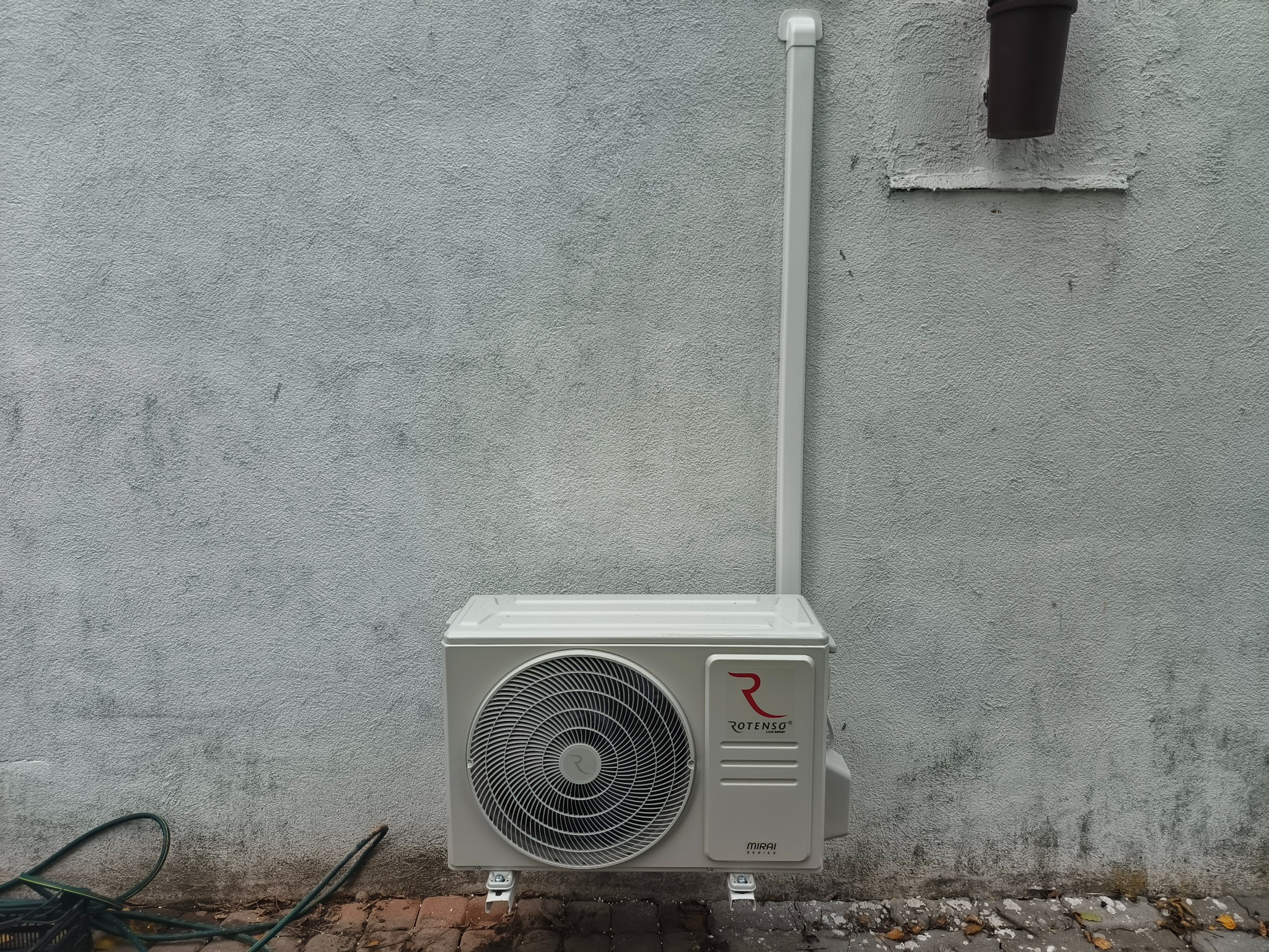 Zamontowana jednostka zewnętrzna klimatyzacji Rotenso MIRAI na szarej, chropowatej ścianie budynku. Widoczna instalacja odprowadzająca skropliny i fragment węża ogrodowego.