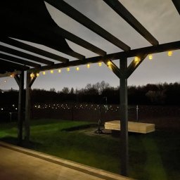 Belka (BLK Woodworking) - Oświetlona pergola z ciemnego drewna, z wiszącymi lampionami, widziana nocą z tarasu. W tle zieleń i delikatne światła w oddali.