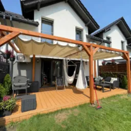 Nowoczesny taras z drewnianą pergolą i meblami ogrodowymi, hamaki wiszące, grill, zadbany trawnik. Słoneczny dzień, widok na biały dom z czarnym dachem.