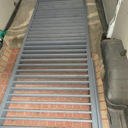 Belka (BLK Woodworking) - Szara, drewniana konstrukcja z listew, prawdopodobnie element balustrady lub schodów, leży wewnątrz pojazdu dostawczego na dywanie.