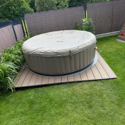 Belka (BLK Woodworking) - Owalna wanna spa z pokrywą, ustawiona na niskim tarasie z desek kompozytowych. Otoczenie: trawnik, ogrodzenie panelowe i zieleń. Ujęcie z poziomu trawnika.
