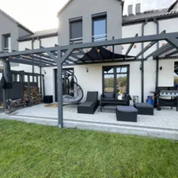 Szara pergola z oświetleniem nad tarasem z meblami ogrodowymi i grillem. Uwagę przyciąga wiszące krzesło i kompozycja drewna na boku. Widok na zadbany trawnik.