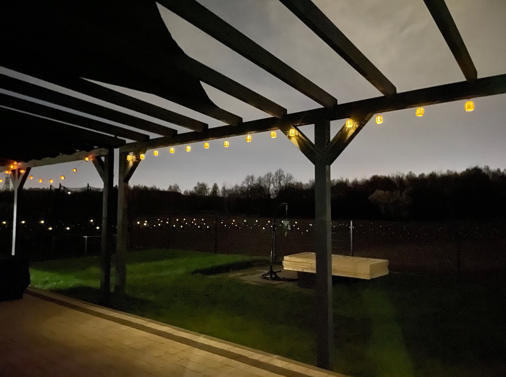Oświetlona pergola z ciemnego drewna, z wiszącymi lampionami, widziana nocą z tarasu. W tle zieleń i delikatne światła w oddali.