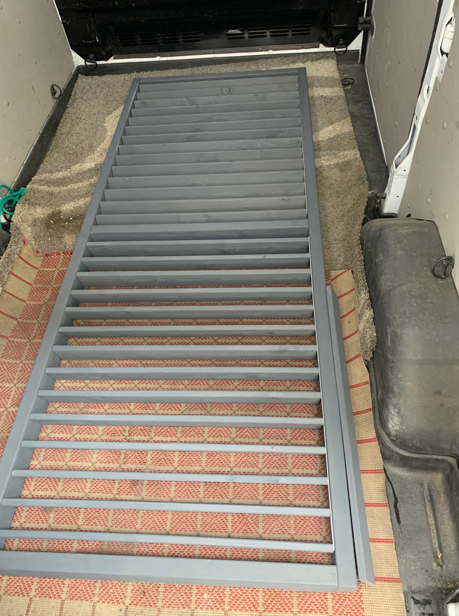 Szara, drewniana konstrukcja z listew, prawdopodobnie element balustrady lub schodów, leży wewnątrz pojazdu dostawczego na dywanie.