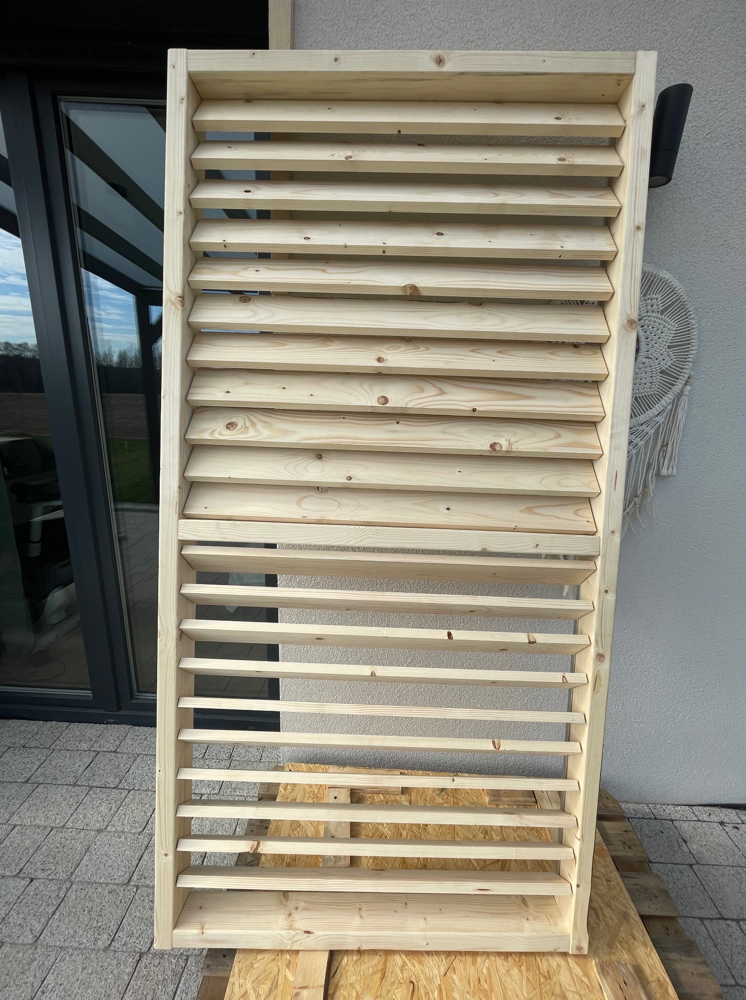 Surowa drewniana konstrukcja z lamelami, oparta o paletę, na tle ściany budynku z oknem i dekoracją. Element do zabudowy balkonu lub tarasu, jasne drewno.