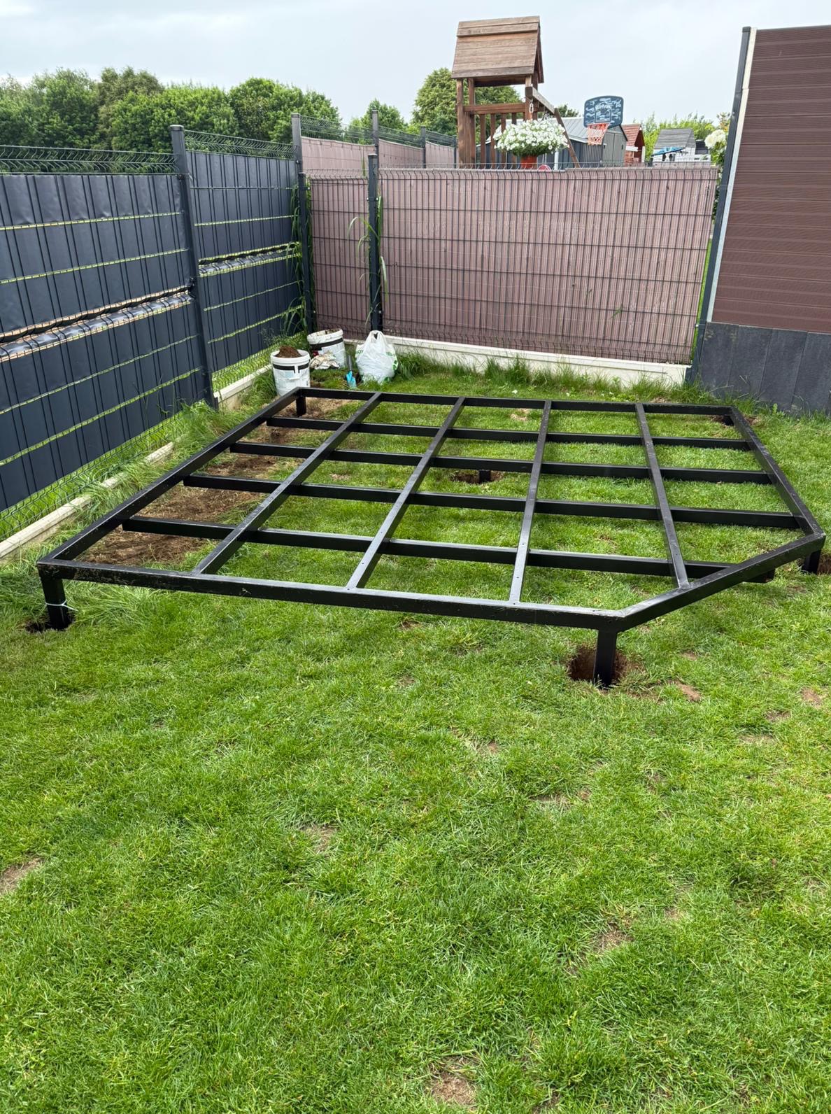 Metalowa konstrukcja tarasu na trawie. W tle ogrodzenie panelowe, worki z ziemią i elementy małej architektury ogrodowej. Widok z góry na szkielet tarasu.