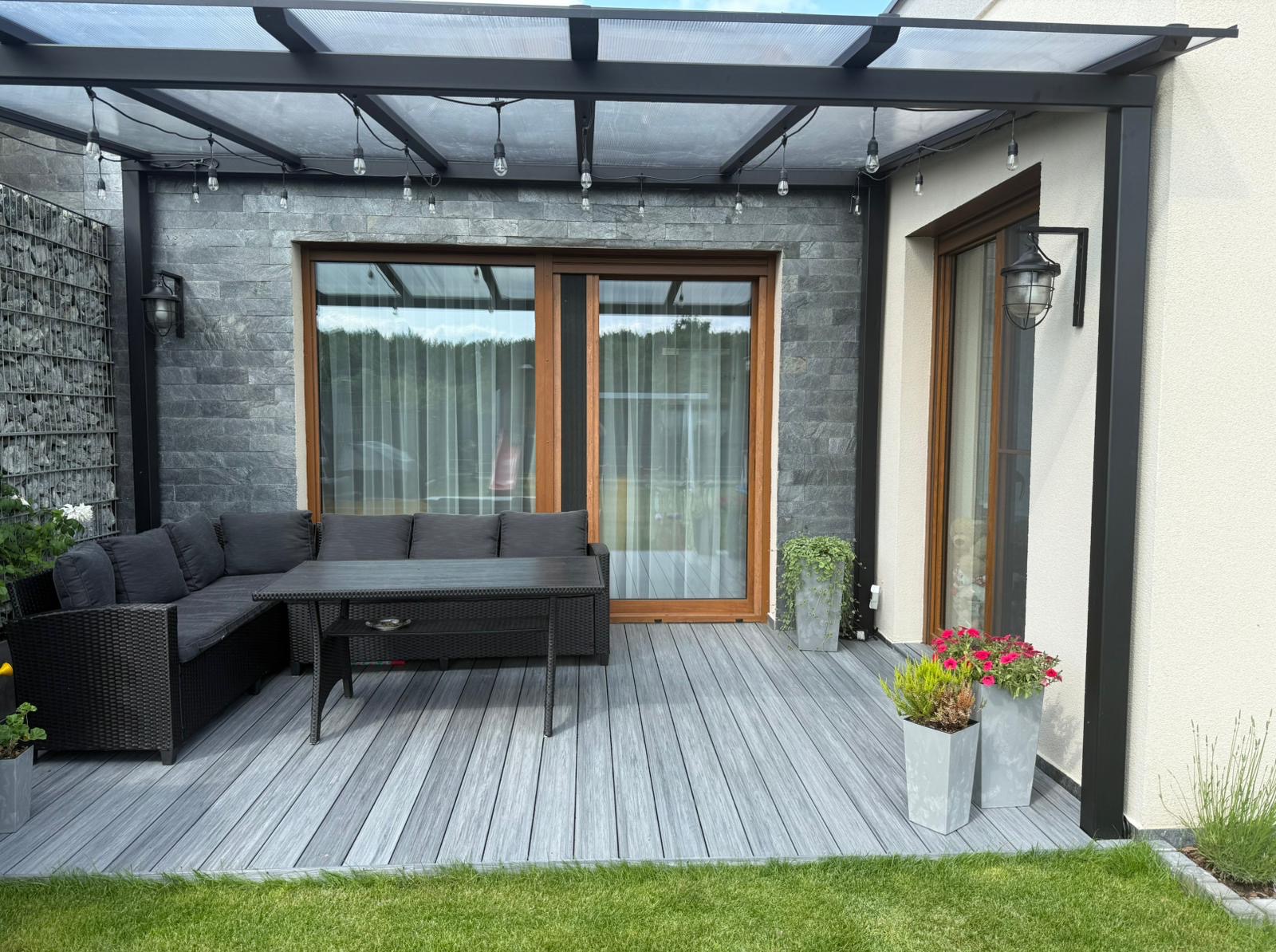 Szary taras kompozytowy z pergolą i meblami ogrodowymi. Widoczne oświetlenie dekoracyjne, rośliny w donicach i fragment trawnika. Nowoczesny design.