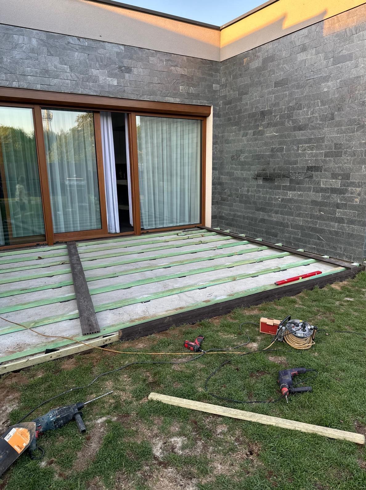 Montaż tarasu: widoczne legary z zieloną izolacją i częściowo ułożone deski w kolorze ciemnego brązu. Na trawie leżą narzędzia: wiertarki, poziomica i przedłużacz.