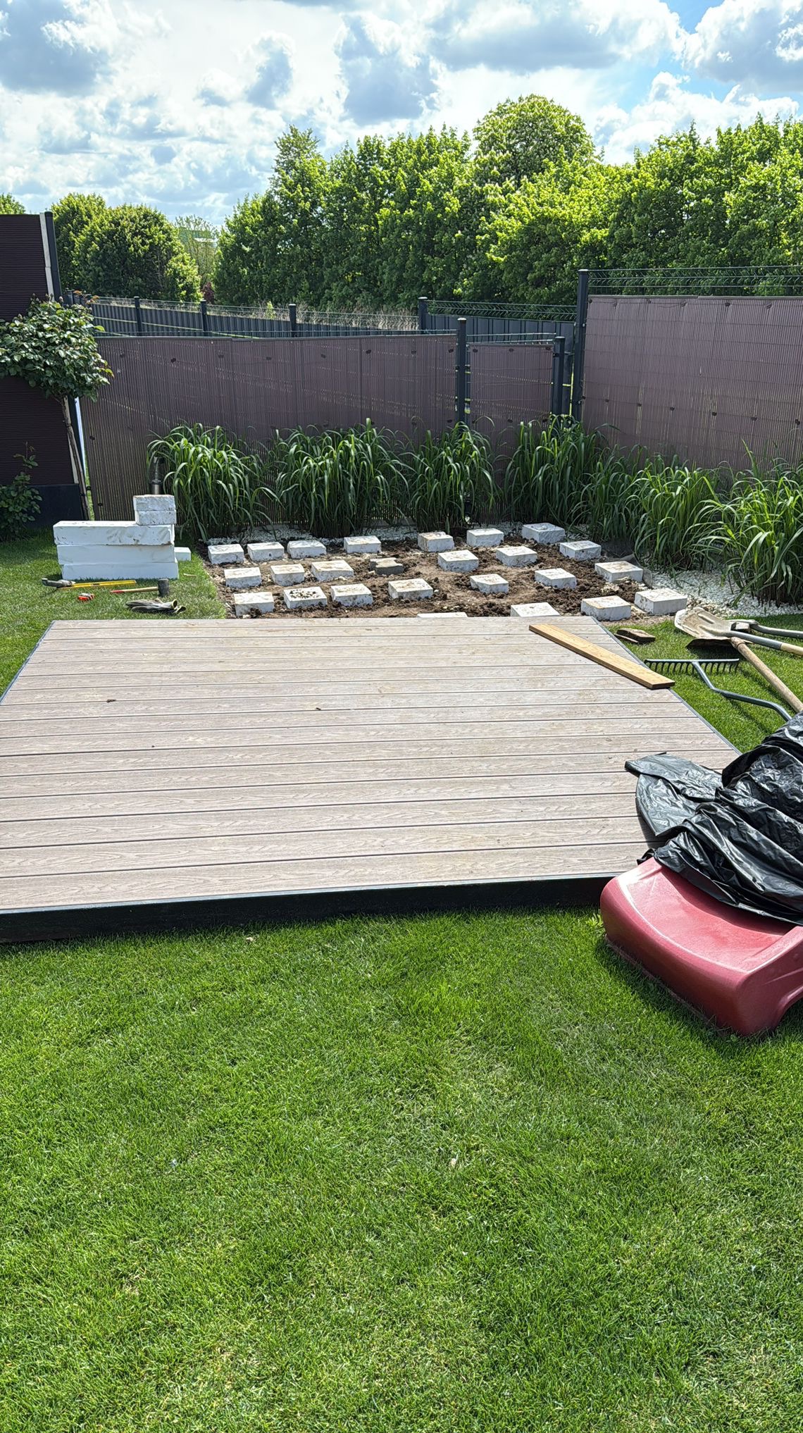 Montaż tarasu z desek kompozytowych w ogrodzie: widoczna część ułożonej podłogi, przygotowane podłoże z kostkami, narzędzia i akcesoria montażowe.