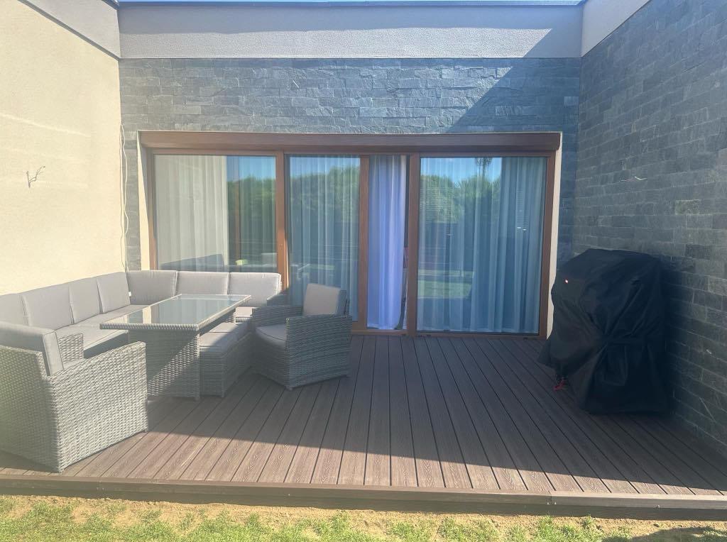 Nowoczesny taras kompozytowy z szarymi meblami ogrodowymi i grillem pod przykryciem, przylegający do budynku z kamienną elewacją i dużymi oknami.