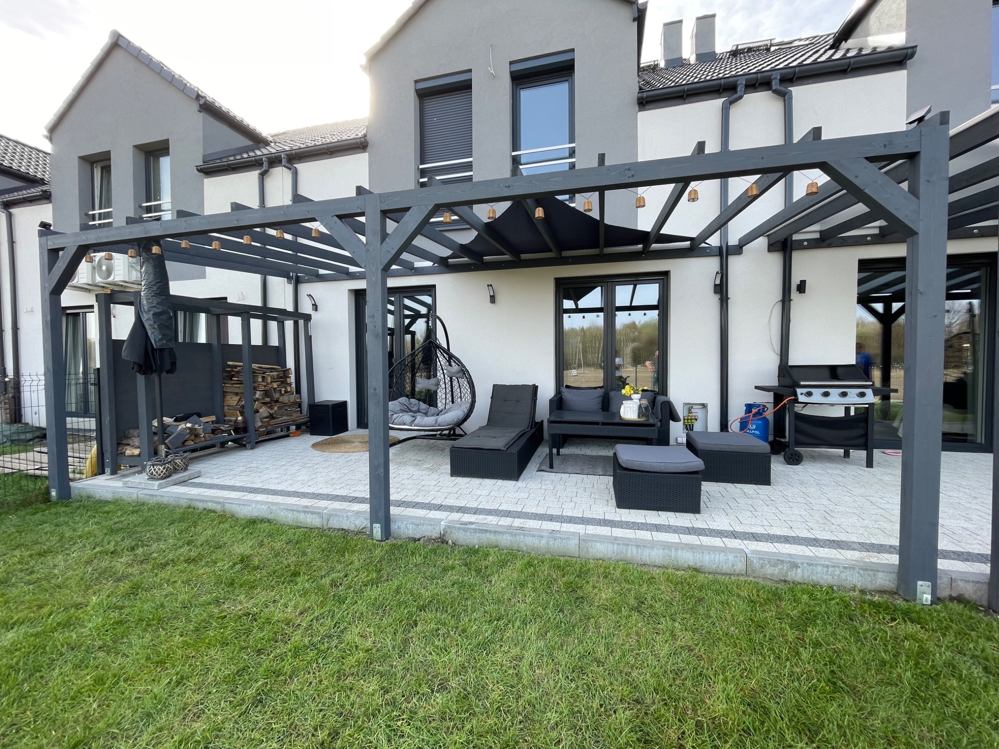 Szara pergola z oświetleniem nad tarasem z meblami ogrodowymi i grillem. Uwagę przyciąga wiszące krzesło i kompozycja drewna na boku. Widok na zadbany trawnik.