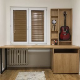 Drewniane biurko z szafką i półkami, na których stoi gitara i zegar. Blat biurka przechodzi nad grzejnikiem. Okno z żaluzjami w tle.