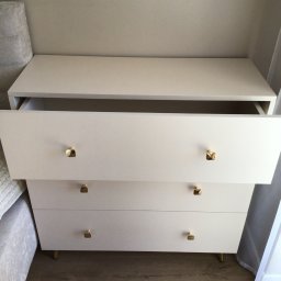 Jasna komoda z trzema szufladami, jedna otwarta, z złotymi uchwytami i nóżkami, ustawiona na drewnianej podłodze, obok łóżka i zasłony.