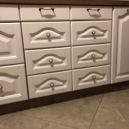 Białe, frezowane fronty szuflad i szafki kuchennej z ciemnobrązowym blatem i cokołem, widoczne uchwyty i gałki w stylu retro, na tle płytek podłogowych.