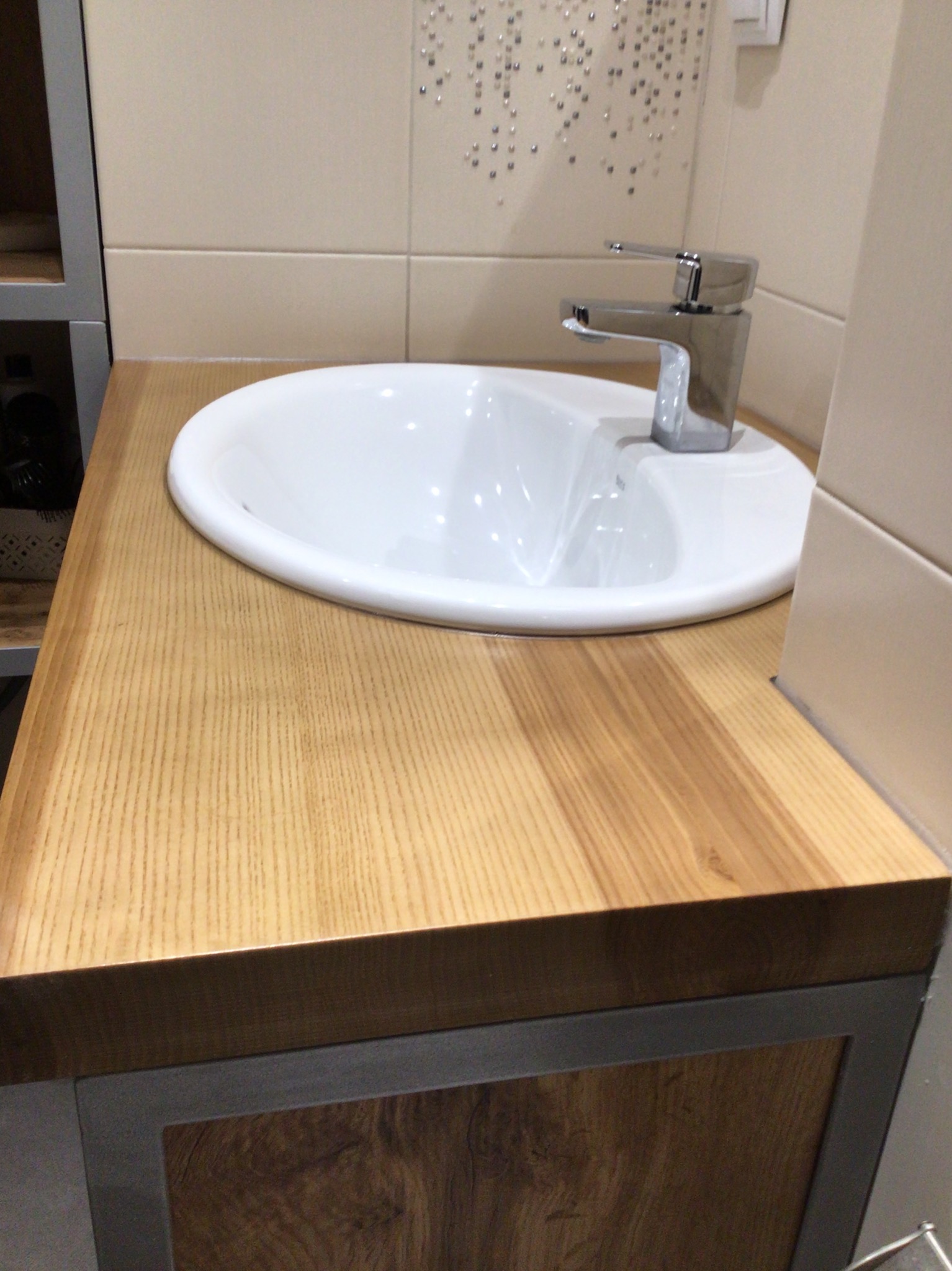 Biała umywalka nablatowa z chromowaną baterią na blacie z jasnego drewna, osadzona na szafce z ciemniejszego drewna. Tło stanowią beżowe płytki z dekoracyjnymi elementami.