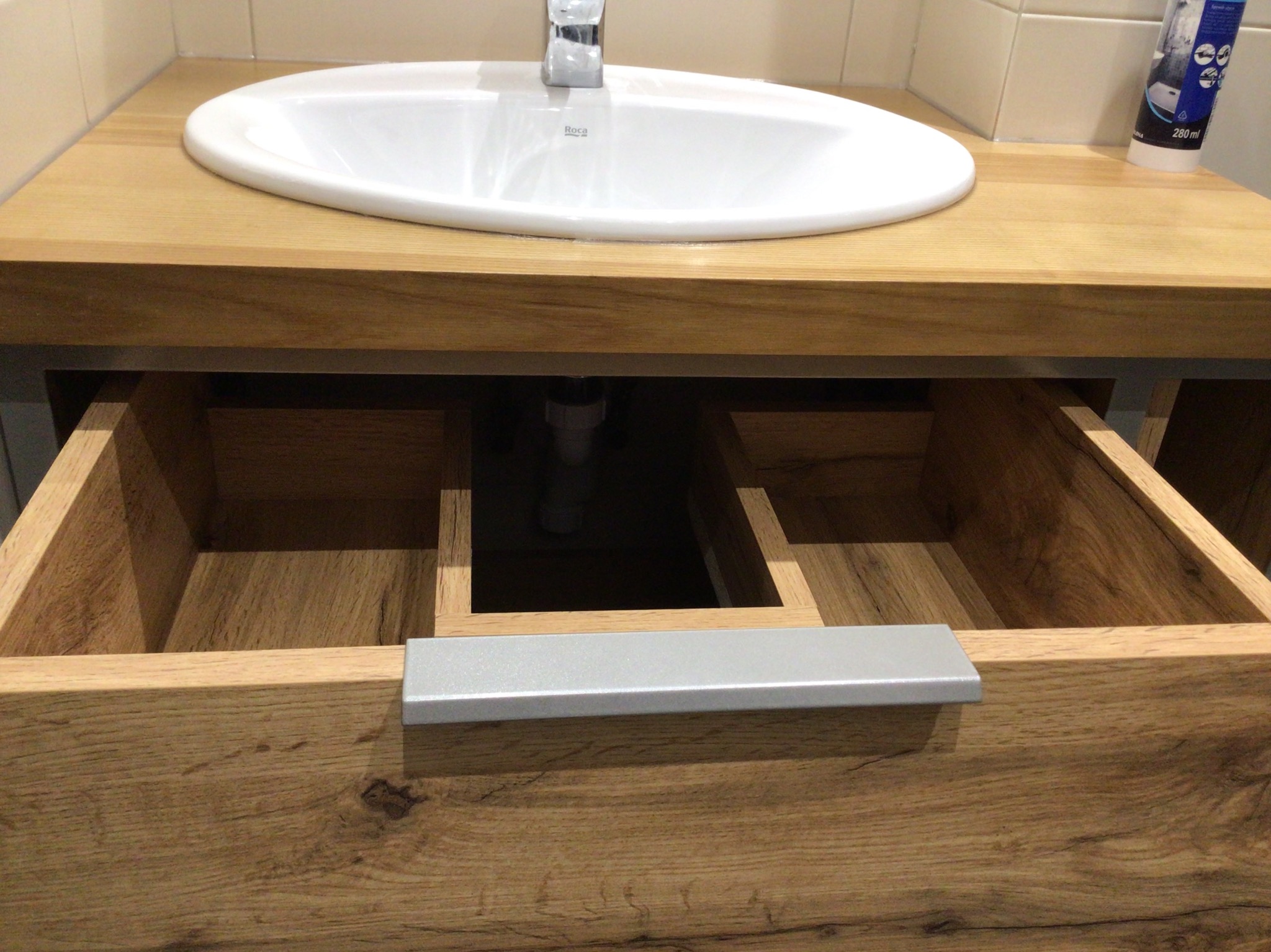 Otwarta szuflada z drewna pod umywalką w łazience, widoczna struktura drewna i metalowy uchwyt. Umywalka owalna, biała, zamontowana na drewnianym blacie.