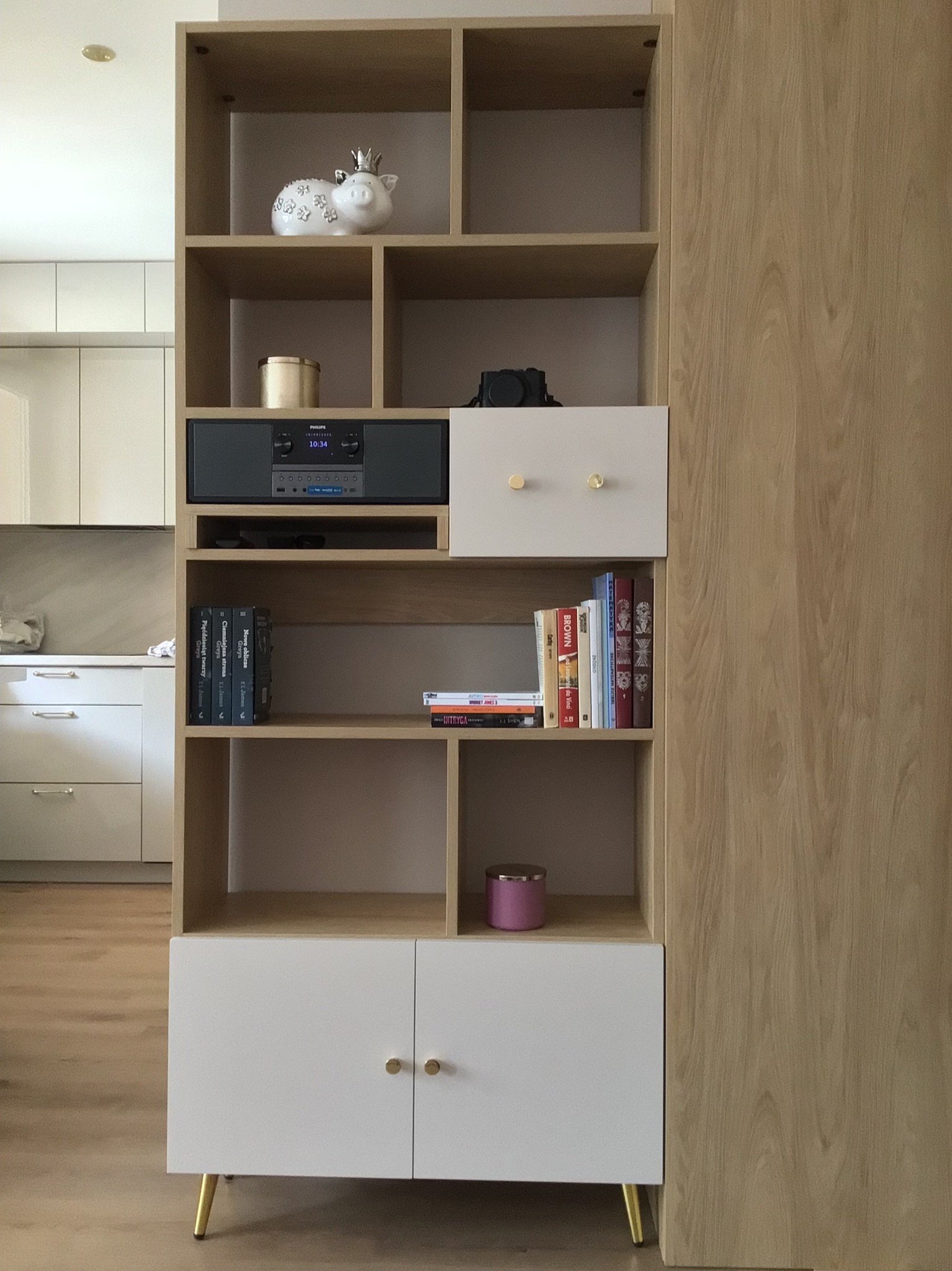 Regał z jasnego drewna z białymi frontami, złotymi nóżkami i gałkami. Na półkach książki, radio, dekoracje. Widoczny fragment kuchni w tle. Ujęcie z bliska.