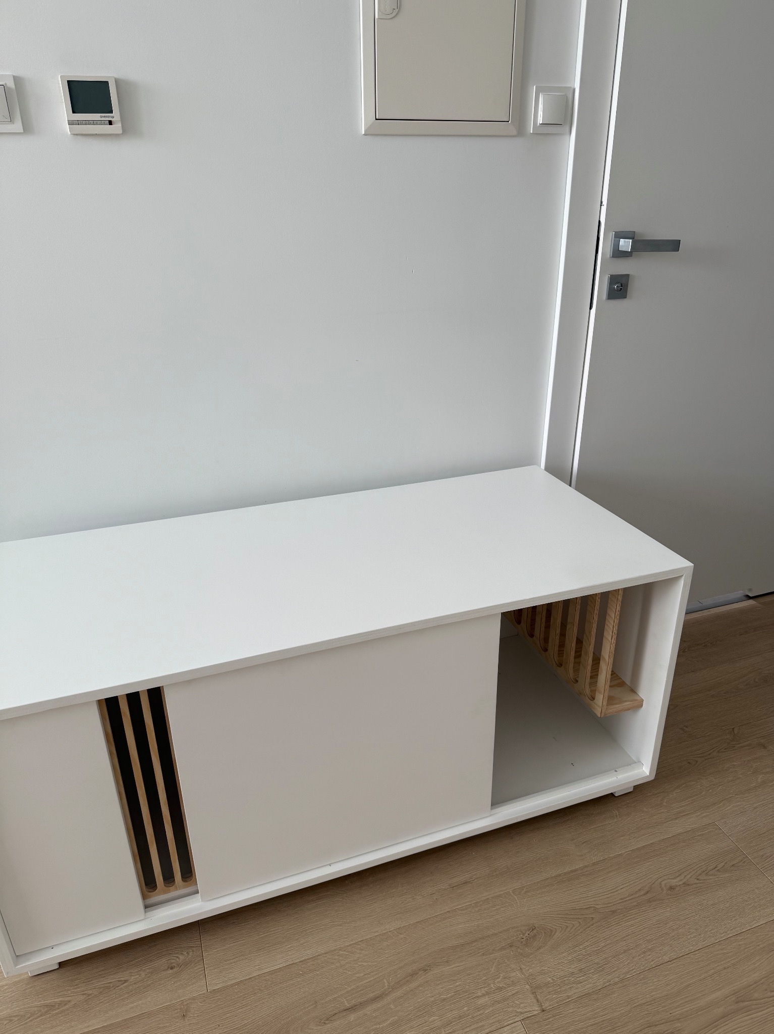 Biała szafka RTV z przesuwnymi drzwiami i drewnianymi akcentami, ustawiona na podłodze w jasnym wnętrzu. Minimalistyczny design, widoczne detale wykończenia.