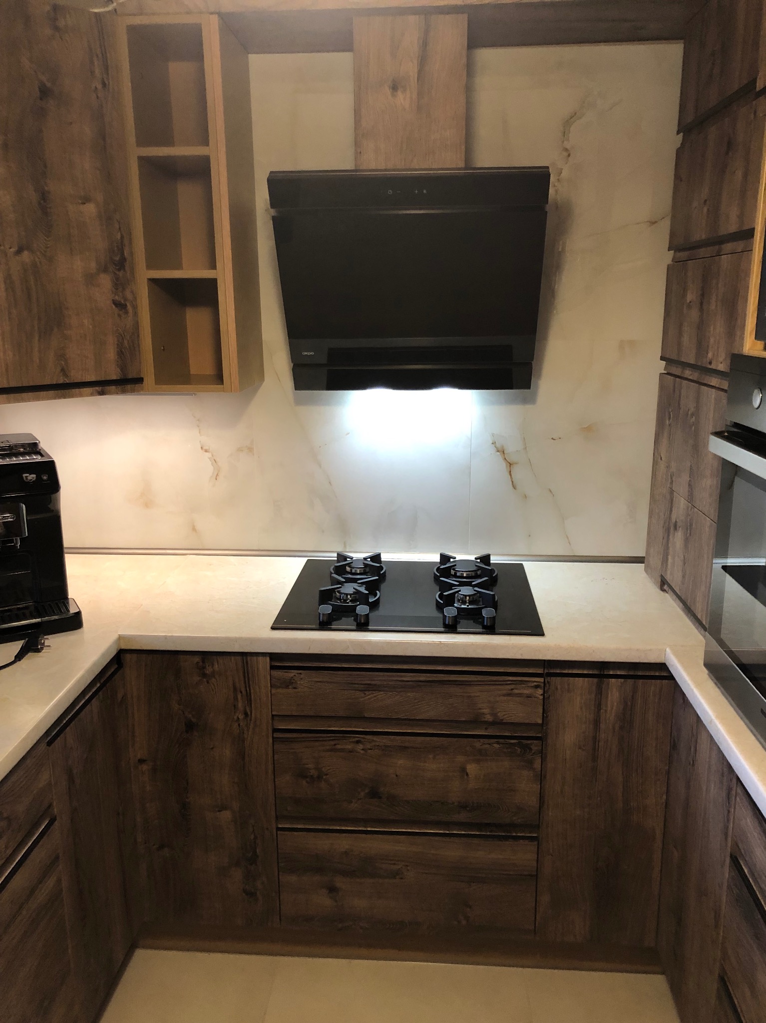 Nowoczesna kuchnia z drewnianymi szafkami, czarną płytą grzewczą i okapem. Blat w jasnym odcieniu marmuru. Widoczny ekspres do kawy i piekarnik.