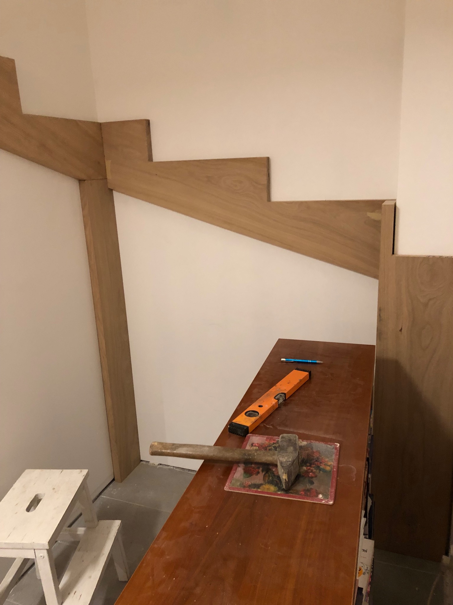 Wnętrze w trakcie montażu drewnianych schodów. Widoczne elementy konstrukcyjne, poziomica, młotek i ołówek na blacie. Etap budowy z surowym wykończeniem ścian.
