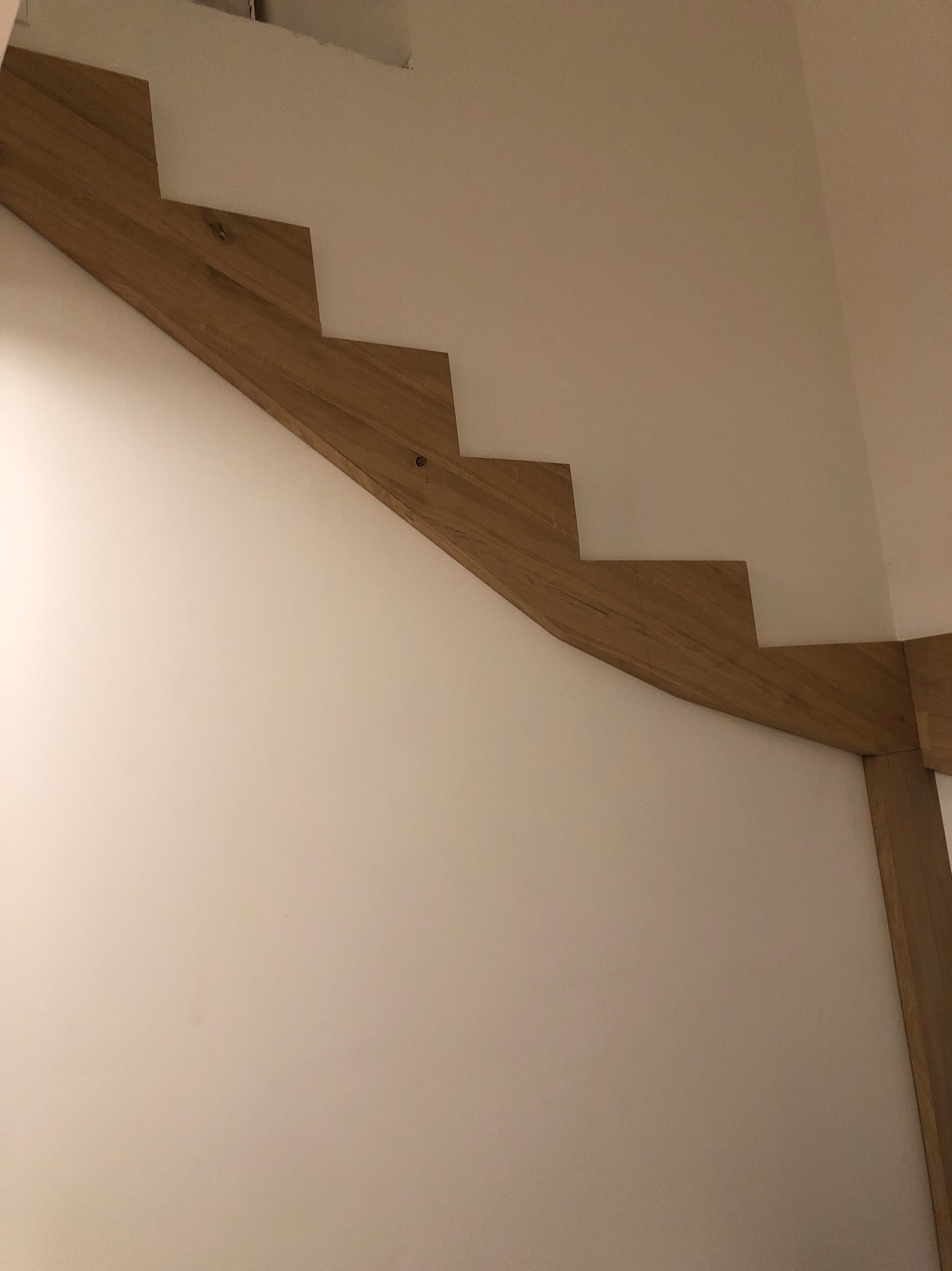 Fragment drewnianych schodów przymocowanych do białej ściany, widoczne stopnie i fragment konstrukcji nośnej. Ujęcie z dołu, podkreślające minimalistyczny design.