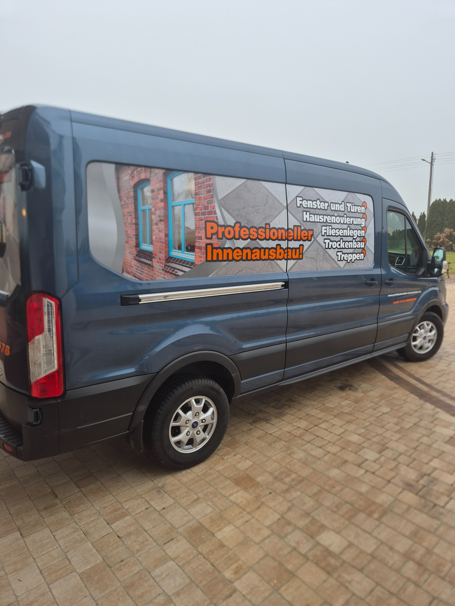 Granatowy bus firmy remontowej z Wijewa, z reklamą usług: okna, drzwi, remonty domów, układanie płytek, zabudowa wnętrz i schody. Widok z boku, na kostce brukowej.
