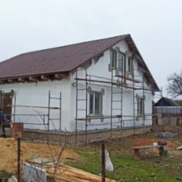 Dom w trakcie ocieplania elewacji, z widocznym rusztowaniem i brązowym dachem. Prace wykończeniowe w toku, widoczne materiały budowlane na posesji.