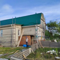 VITALII LAKHAI - Dom z nowym, zielonym dachem z blachy trapezowej, otoczony rusztowaniami. Na pierwszym planie gruz i spacerujący mężczyzna. W tle widać stado kaczek.