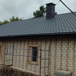 VITALII LAKHAI - Szara blacha trapezowa na dachu domu z kominem. Widoczne rusztowania i przygotowana elewacja z drewnianymi listwami. Pochmurne niebo w tle.