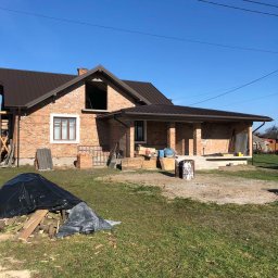 VITALII LAKHAI - Ceglany dom z brązowym dachem w trakcie budowy. Widoczne rusztowania, materiały budowlane i niezadaszony taras. Błękitne niebo w tle.