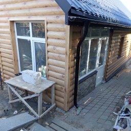 VITALII LAKHAI - Dom z drewnianą elewacją w trakcie prac wykończeniowych, widoczny fragment dachu z orynnowaniem. Stolik roboczy z bloczkiem przed oknem. Nieukończone schody.