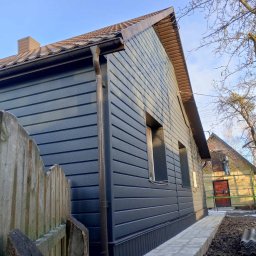 VITALII LAKHAI - Elewacja domu z szarą, poziomą okładziną i brązowym dachem. Widoczne rynny, okna i fragment ogrodzenia. W tle drugi budynek w trakcie prac elewacyjnych.