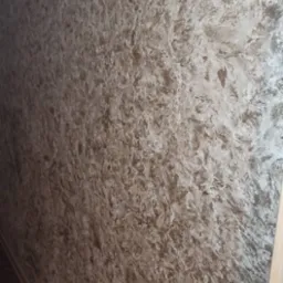 Ściana z dekoracyjnym tynkiem w odcieniach beżu i bieli, wykonana z efektem marmuru. Widoczna faktura i wzór na ścianie. Po prawej stronie fragment futryny drzwi.