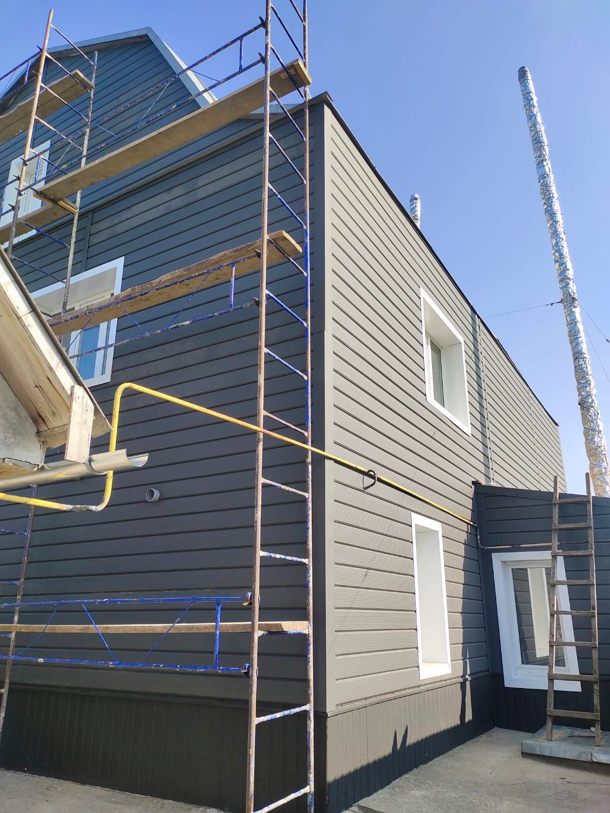 Szare panele elewacyjne na budynku, widoczne rusztowania, rury gazowe i komin. Ujęcie z dołu podkreśla wysokość budynku i etap prac wykończeniowych.