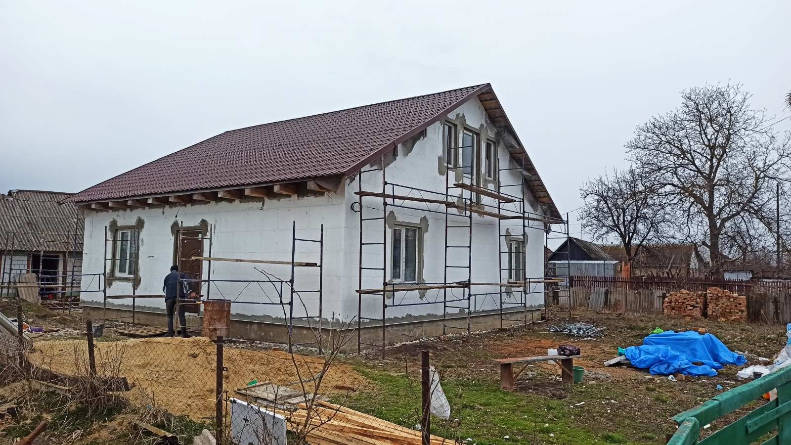 Dom w trakcie ocieplania elewacji, z widocznym rusztowaniem i brązowym dachem. Prace wykończeniowe w toku, widoczne materiały budowlane na posesji.