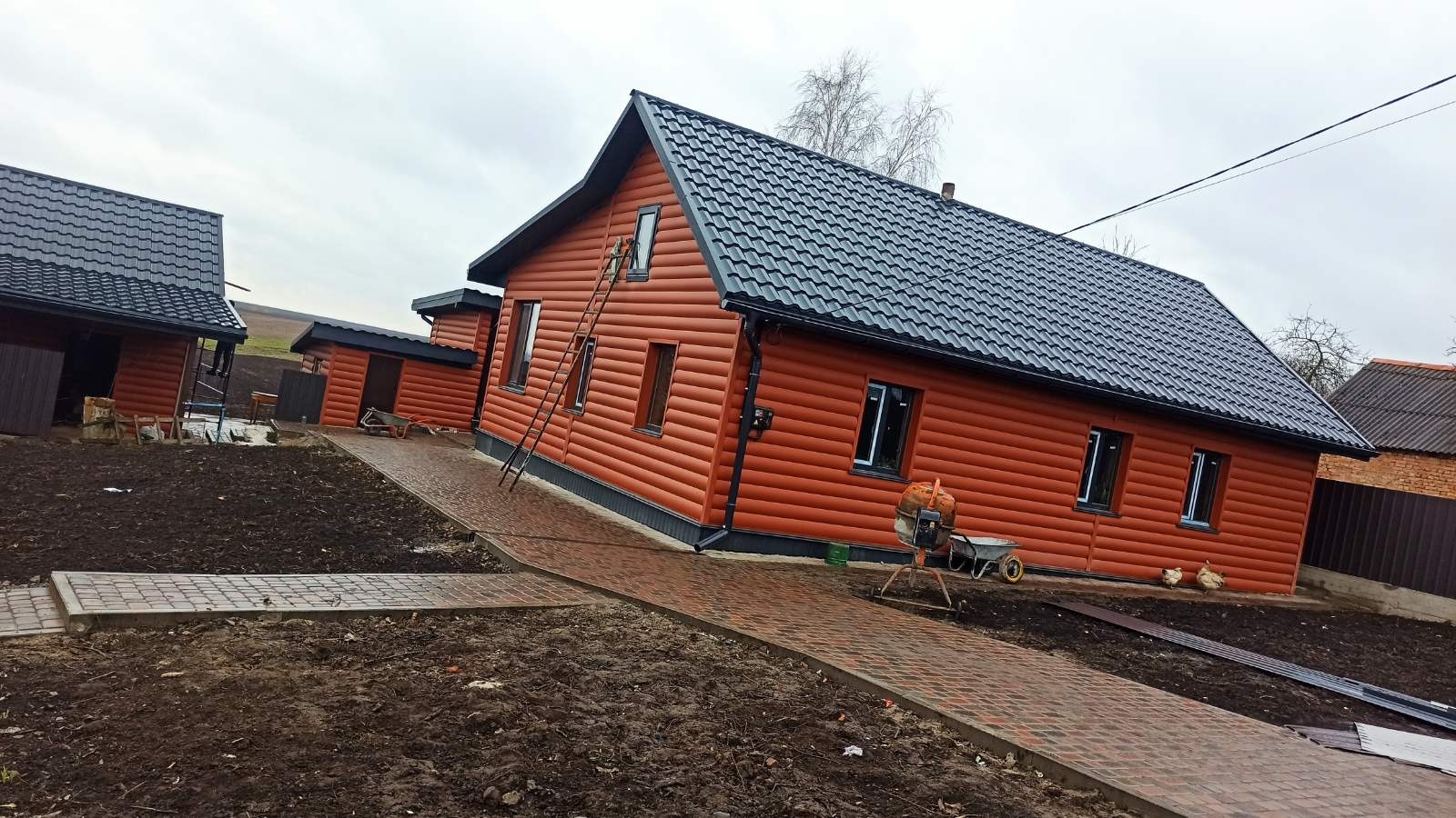 Nowy, ciemny dach z blachy trapezowej na brązowym domu z sidingu, z drabiną opartą o ścianę. Na podwórku widoczny chodnik z kostki i betoniarka.