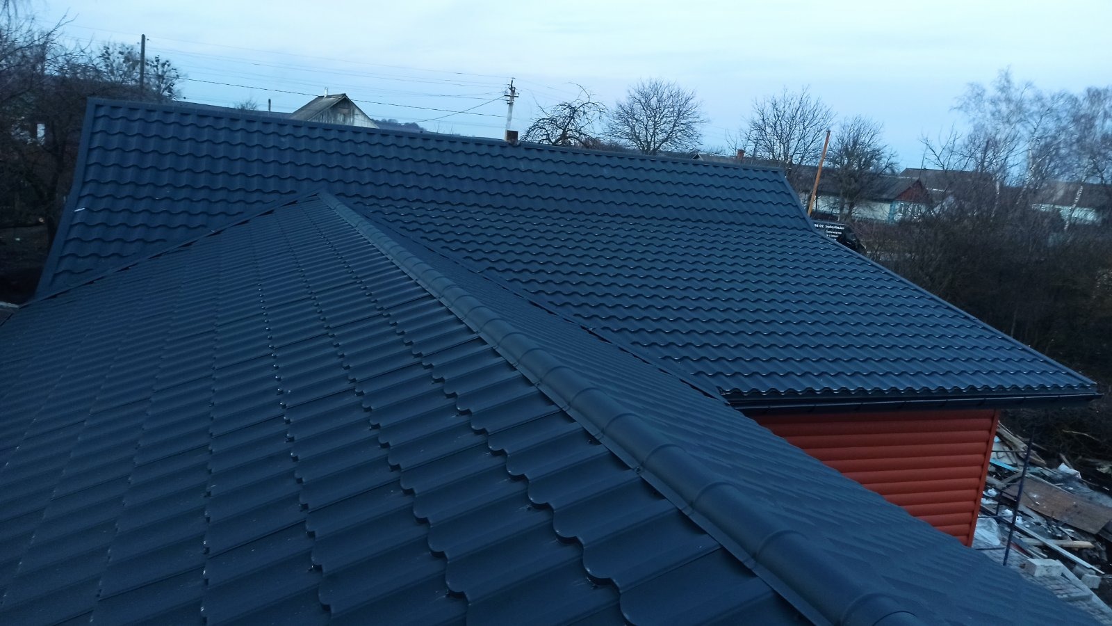 Granatowy dach z blachodachówki modułowej, widoczny z góry, z fragmentem elewacji w kolorze ceglastym. Tło stanowi zabudowa jednorodzinna i drzewa.