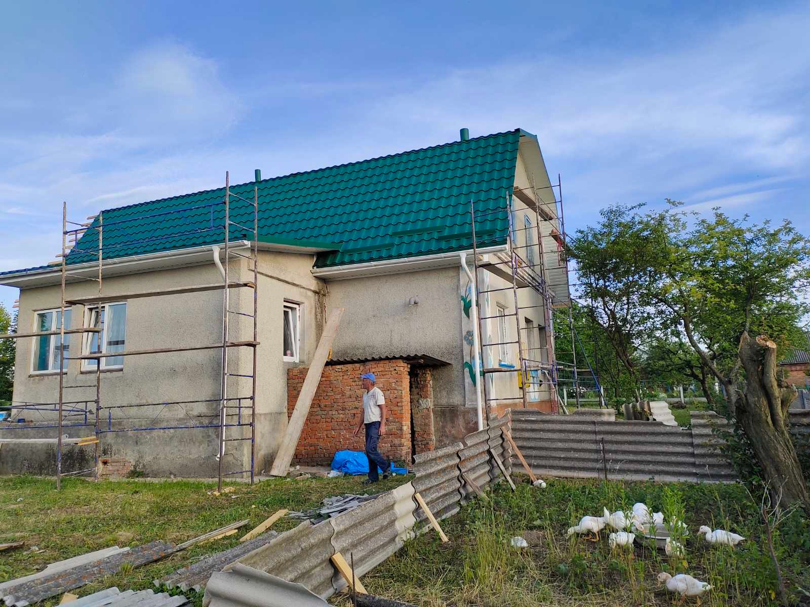 Dom z nowym, zielonym dachem z blachy trapezowej, otoczony rusztowaniami. Na pierwszym planie gruz i spacerujący mężczyzna. W tle widać stado kaczek.