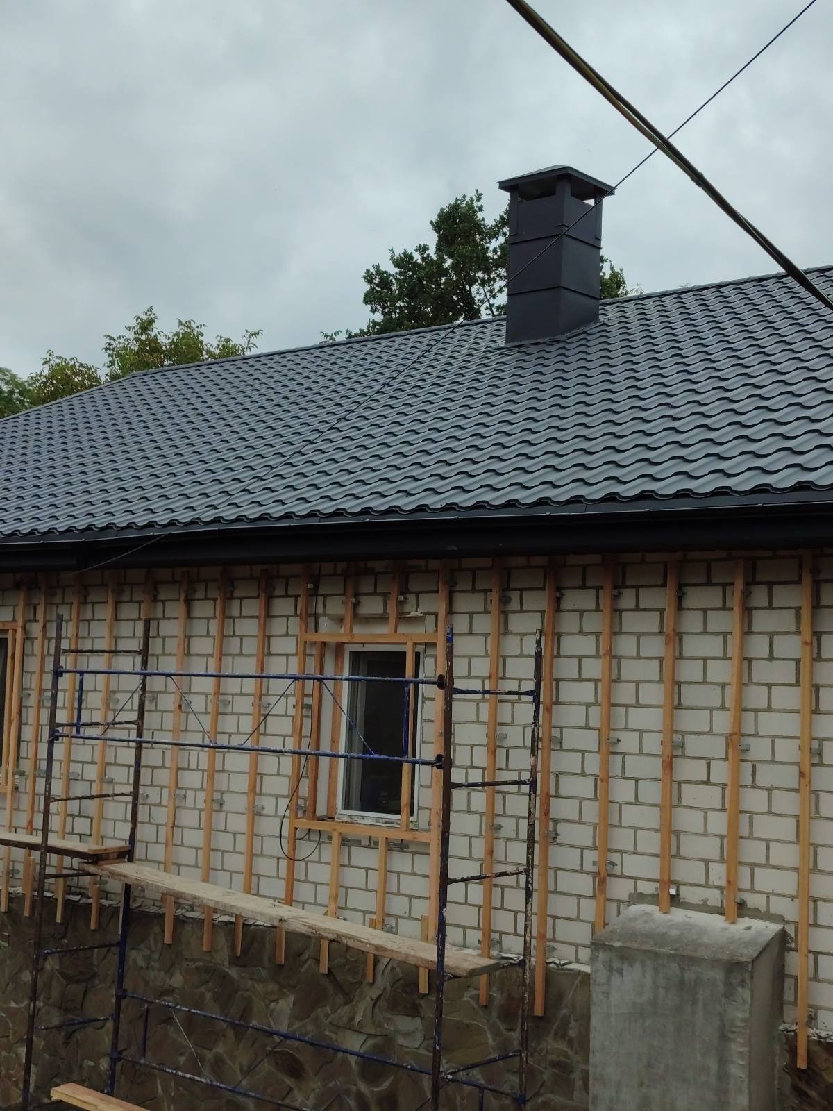Szara blacha trapezowa na dachu domu z kominem. Widoczne rusztowania i przygotowana elewacja z drewnianymi listwami. Pochmurne niebo w tle.