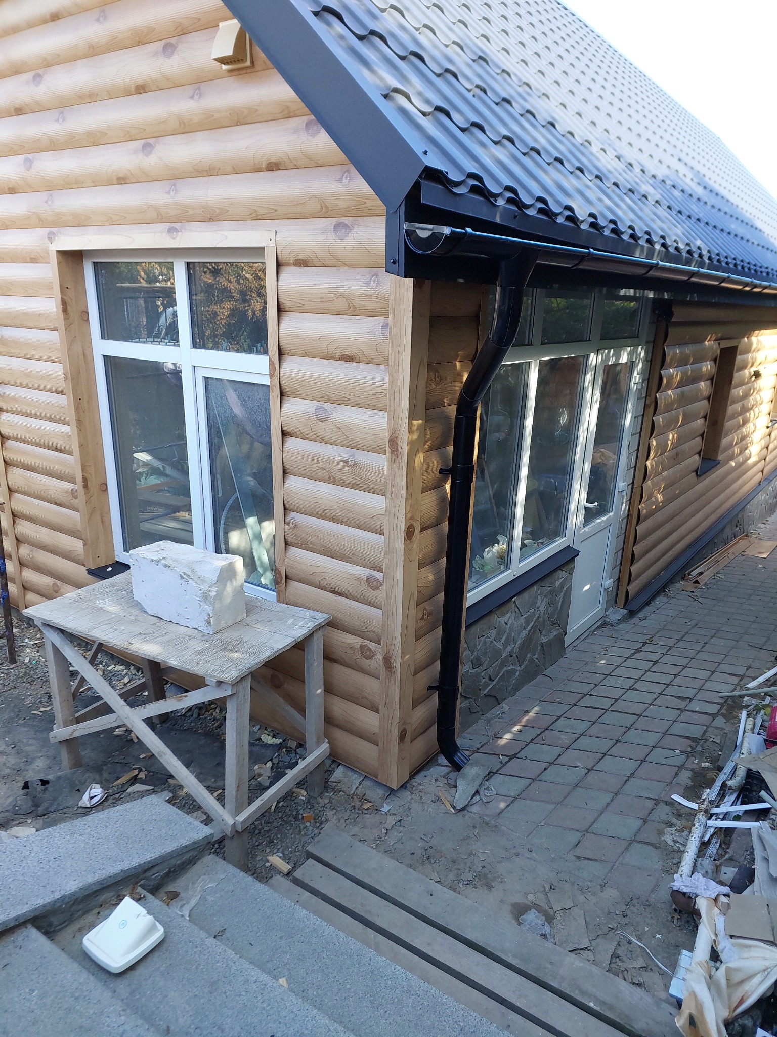Dom z drewnianą elewacją w trakcie prac wykończeniowych, widoczny fragment dachu z orynnowaniem. Stolik roboczy z bloczkiem przed oknem. Nieukończone schody.