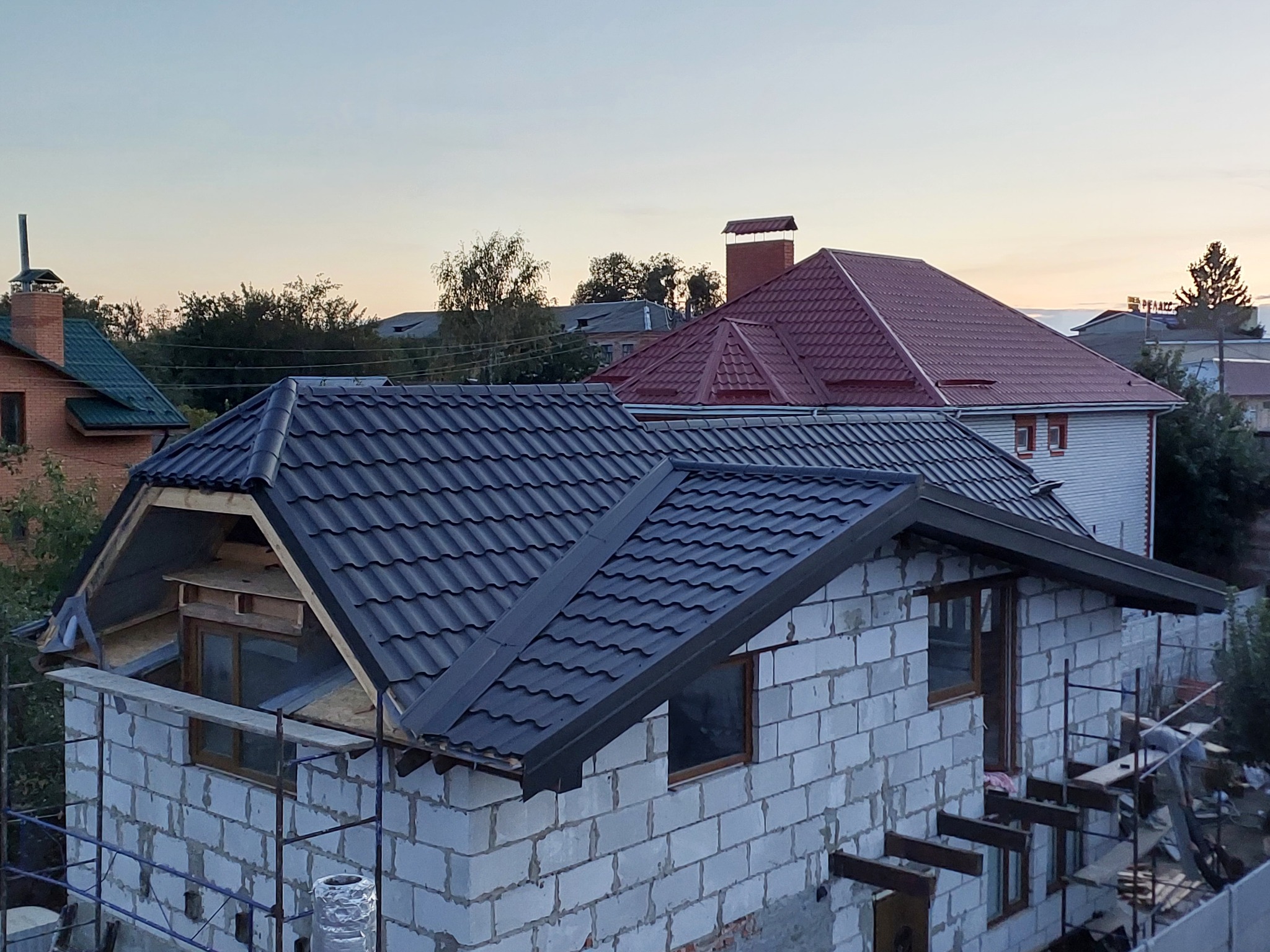 Dom w trakcie budowy z szarym dachem z blachy trapezowej. Widoczne rusztowania i surowy stan ścian z białego pustaka. W tle dom z czerwonym dachem.