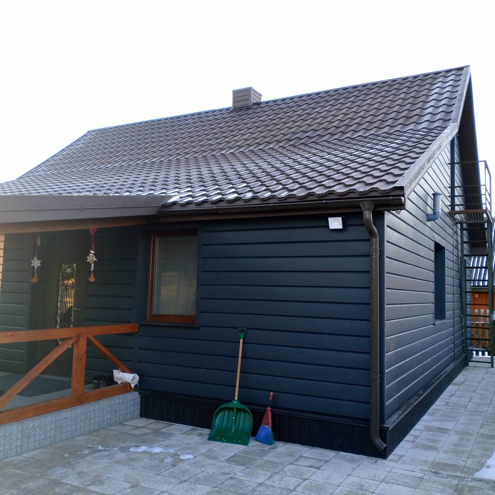 Dom z ciemną elewacją i brązowym dachem z blachodachówki. Widoczne rynny, okno, schody zewnętrzne i narzędzia do odśnieżania.