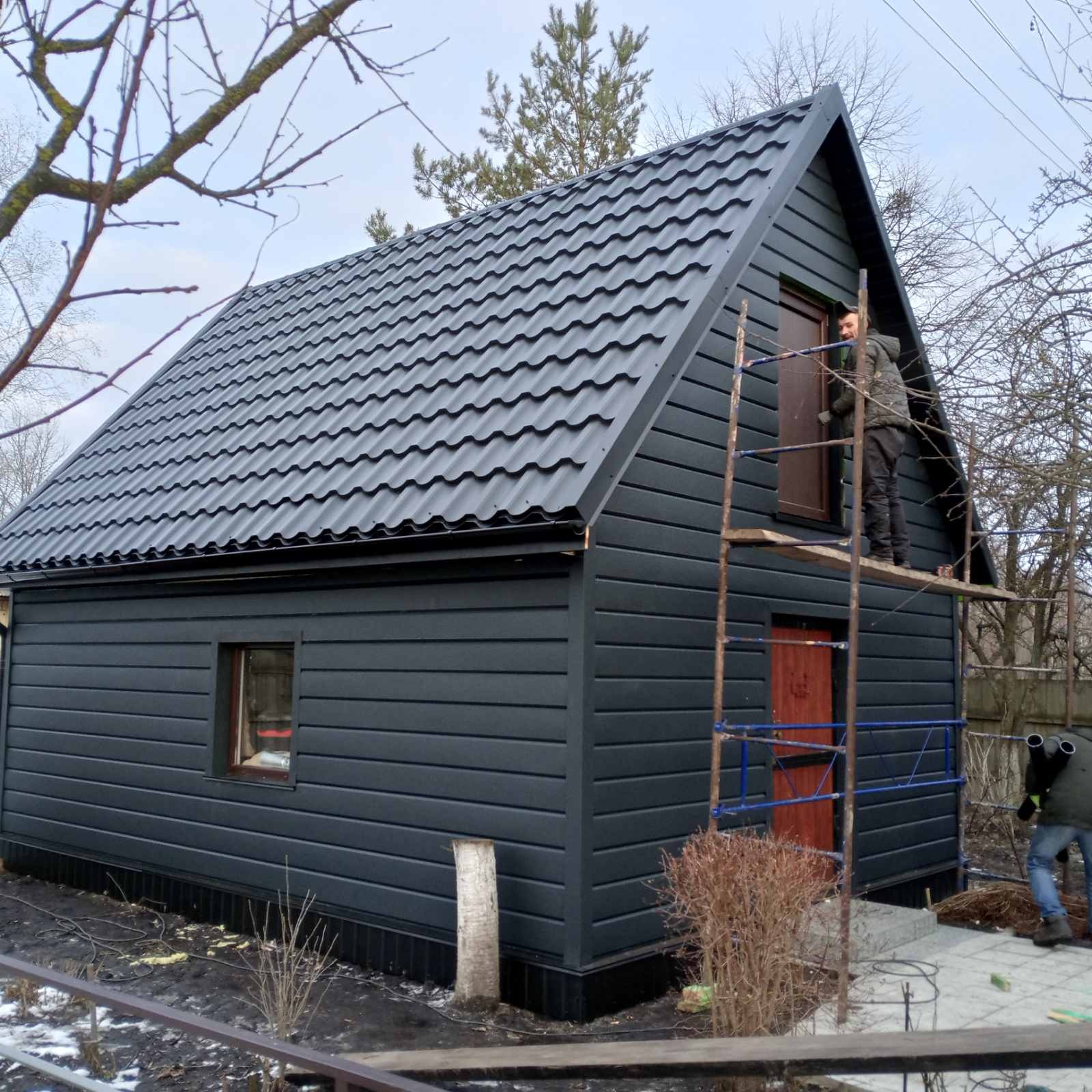 Czarny, nowo wykończony dom z dwuspadowym dachem pokrytym blachodachówką. Pracownik na rusztowaniu montuje okno. Elewacja z paneli. Widok z zewnątrz.