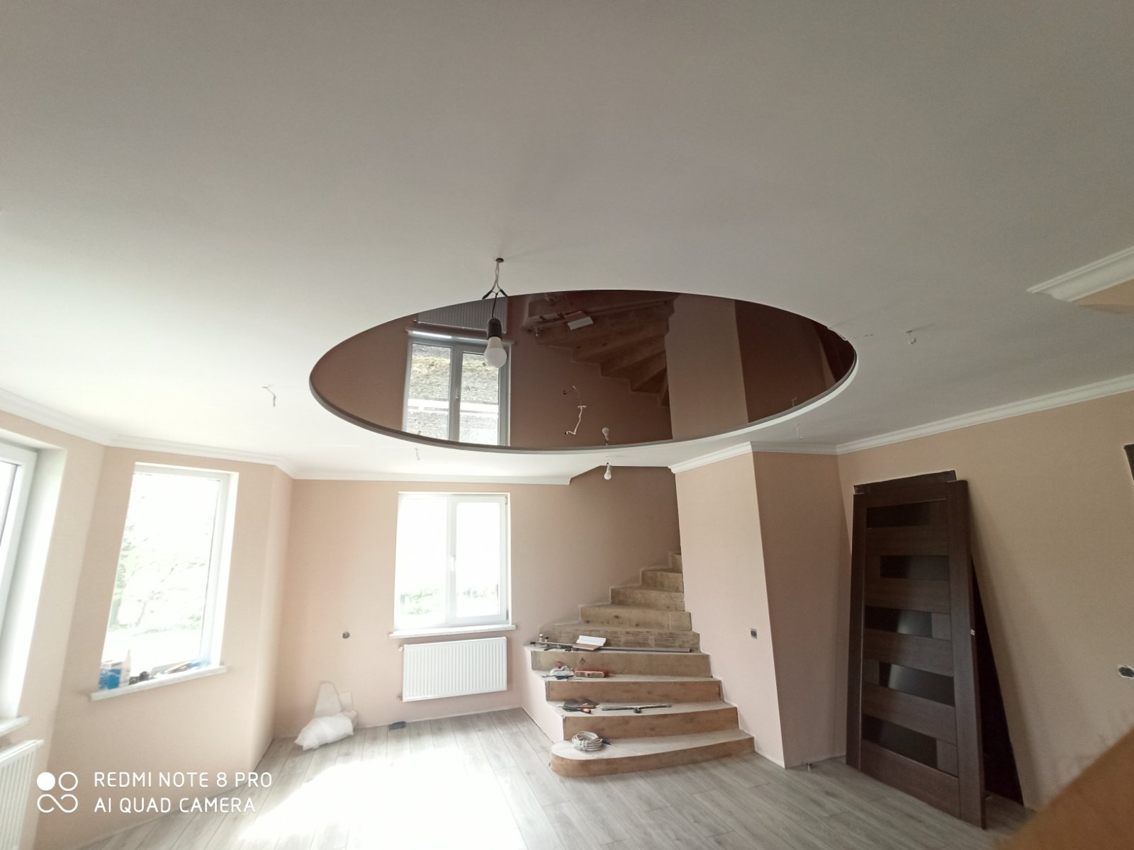 Wnętrze w trakcie wykańczania: sufit z okrągłym lustrem, schody w stanie surowym, częściowo zamontowane drzwi i jasne ściany. Widoczne narzędzia i materiały budowlane.