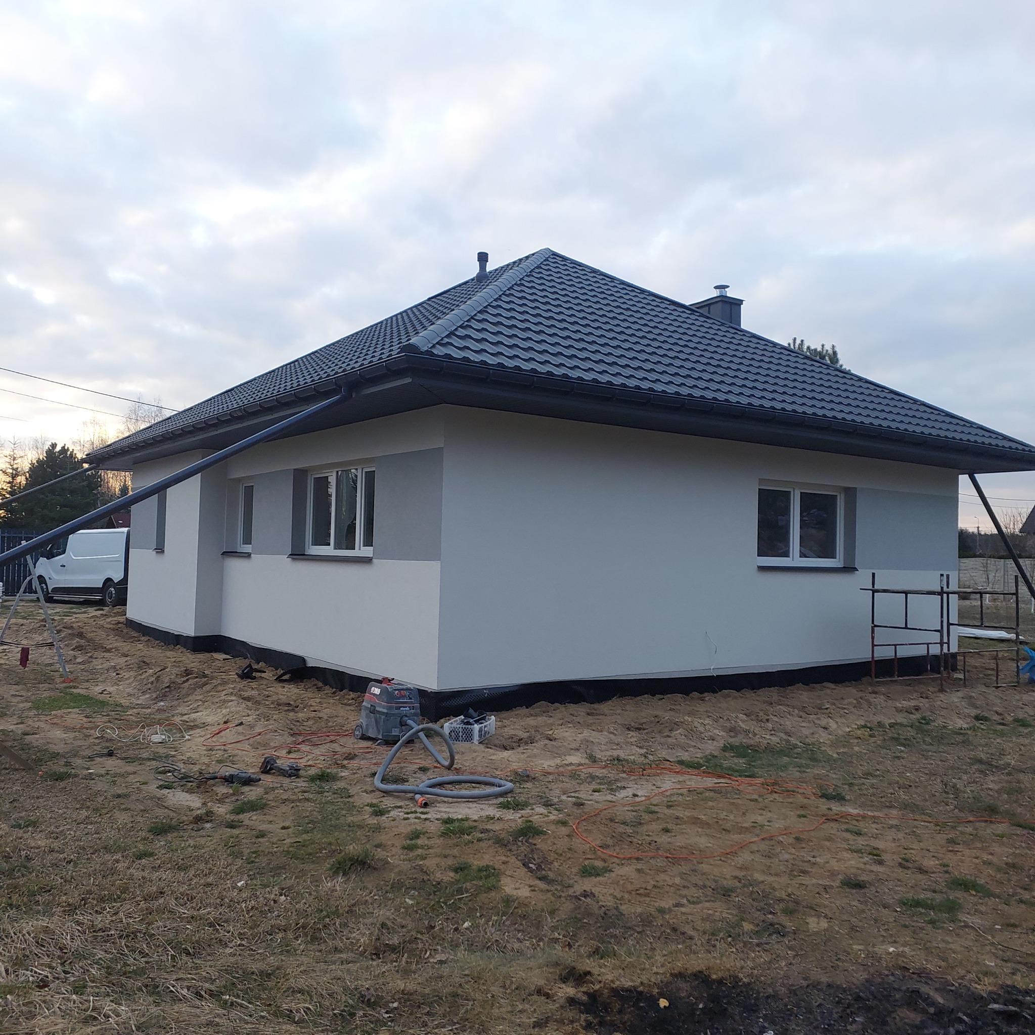 Nowoczesny dom jednorodzinny z szarą dachówką, otynkowany na biało i szaro, z widocznymi narzędziami na posesji. Ujęcie z boku, przy pochmurnym niebie.