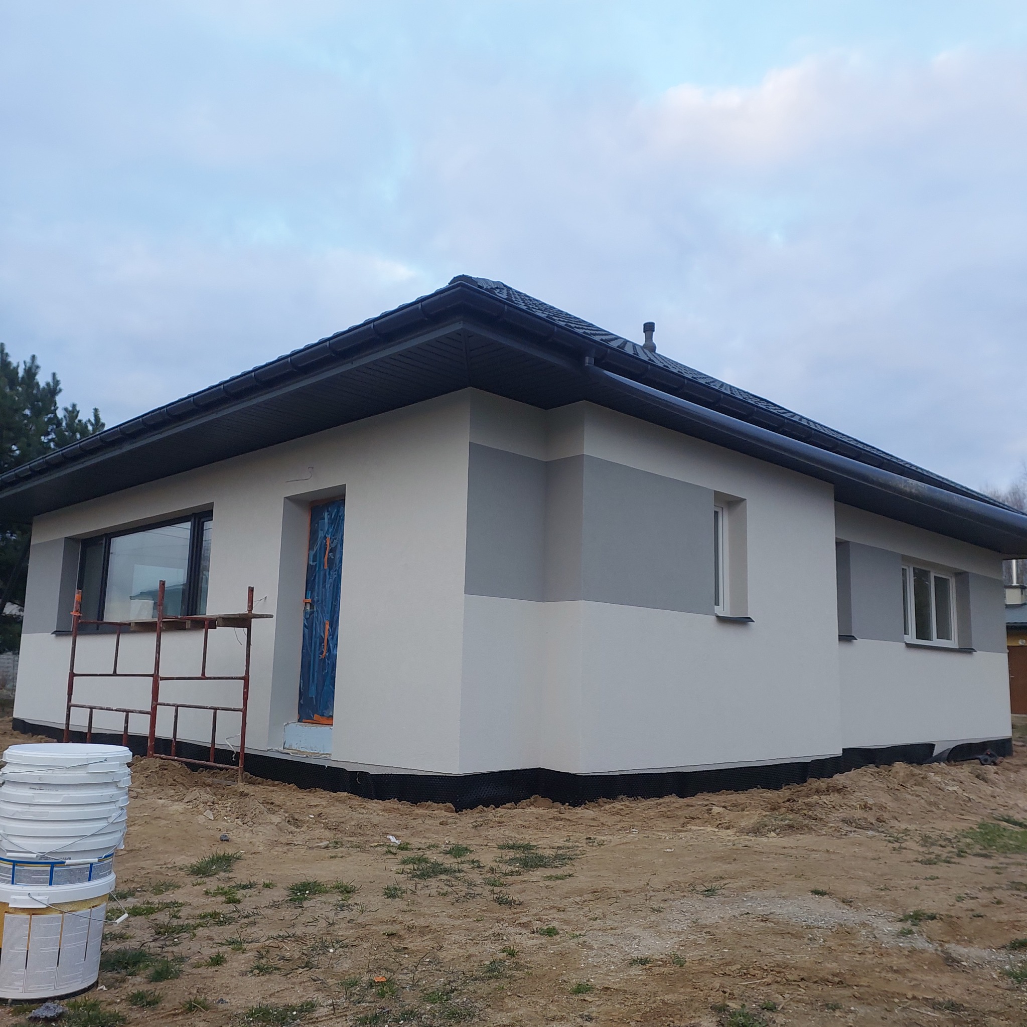 Wykończony dom jednorodzinny z szarą elewacją w pasy, ciemny dach i rusztowanie przy oknie. Ukończone ocieplanie elewacji z widocznymi detalami.