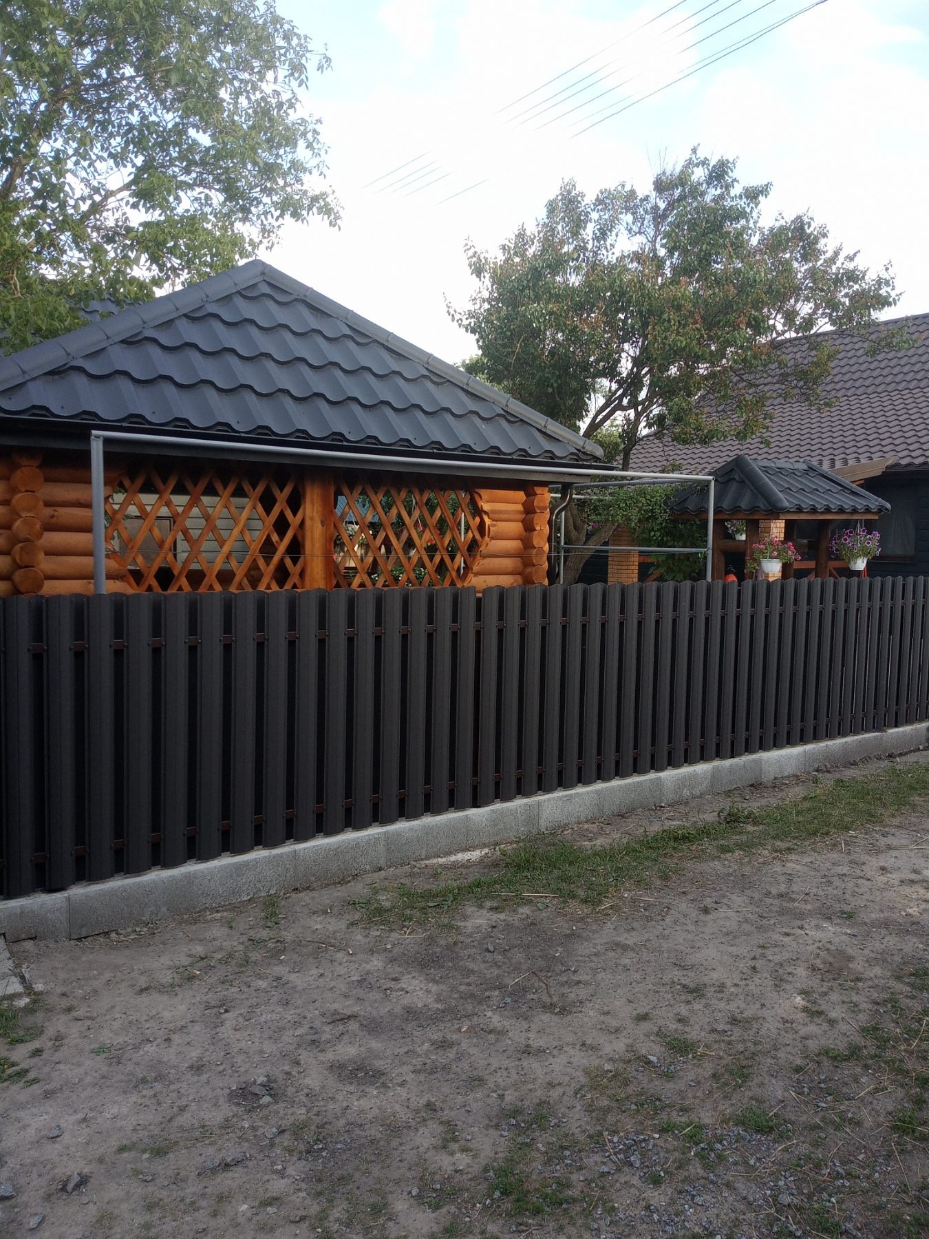 Drewniana altana z czarnym dachem, widoczna przez szary, metalowy płot na betonowej podmurówce. W tle dom z podobnym dachem i roślinność.