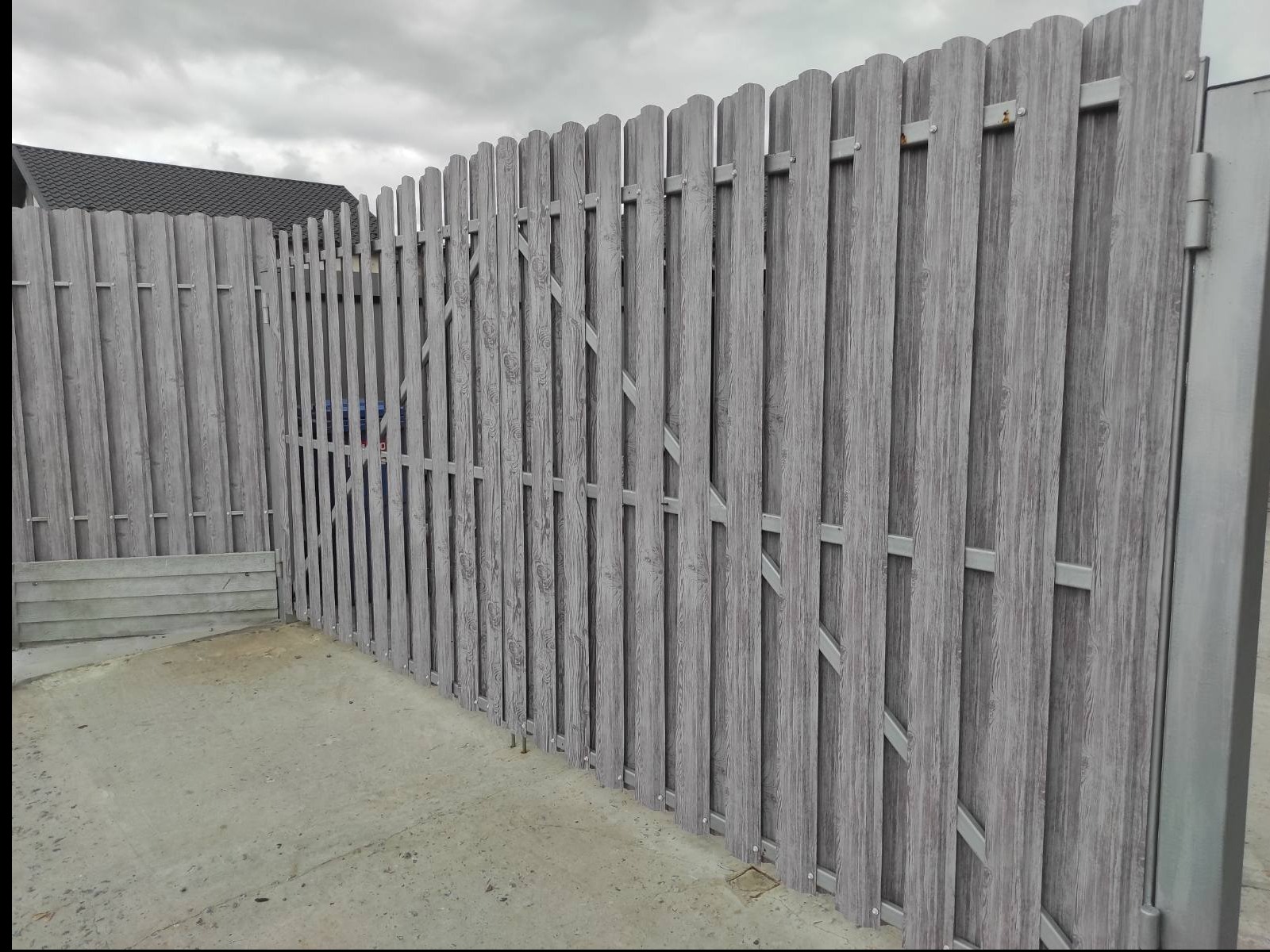 Szara, drewnopodobna, pionowa sztacheta ogrodzeniowa z bramką, widoczna z bliska w Warszawie, na betonowym podłożu, pod pochmurnym niebem.