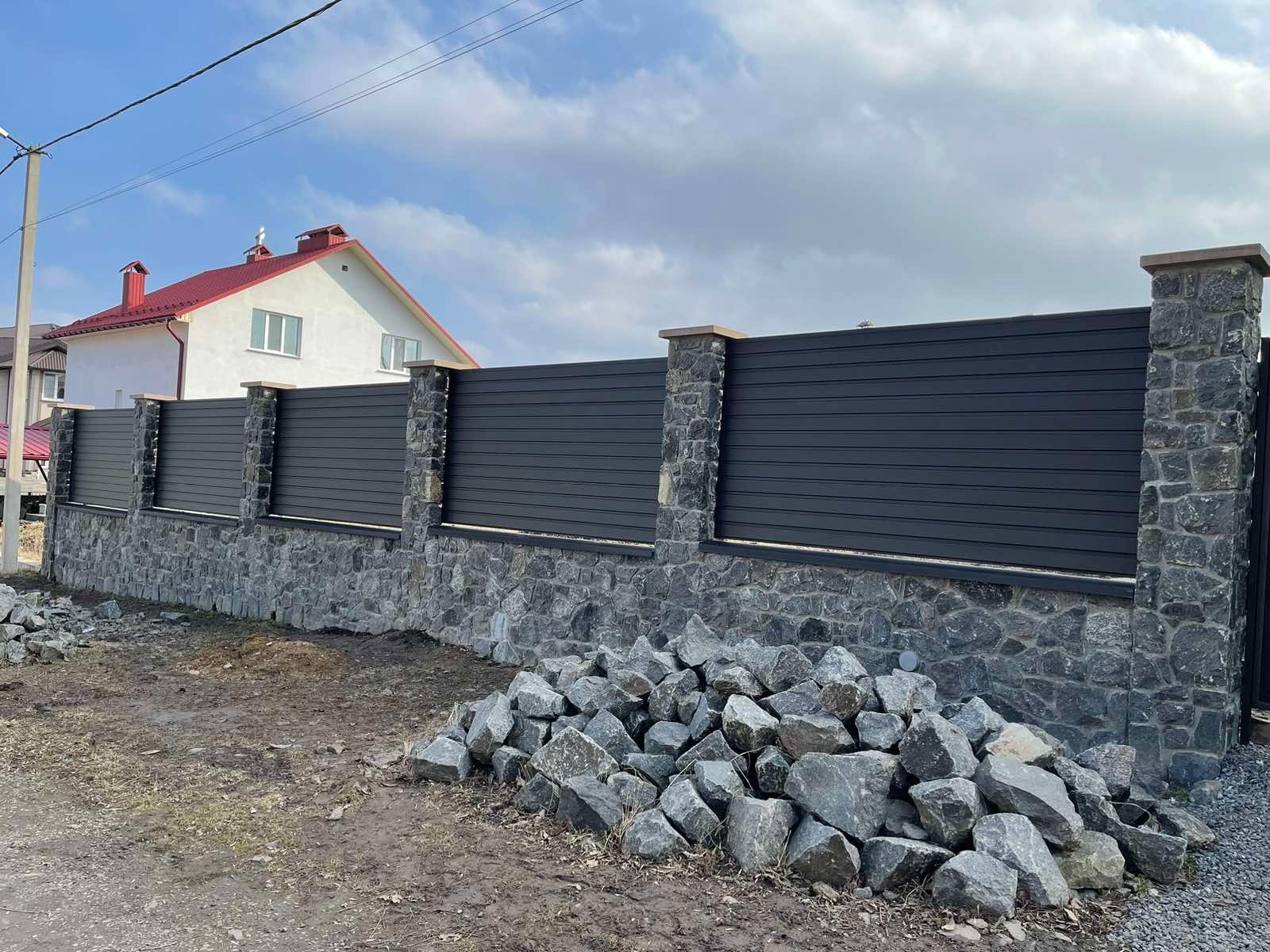 Nowoczesne ogrodzenie z poziomych, grafitowych paneli i kamiennych słupków na tle domu z czerwonym dachem, z stertą kamieni u podstawy.